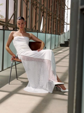 Model wearing a white slip dress with a sheer panel seated on a chair on a modern outdoor terrace