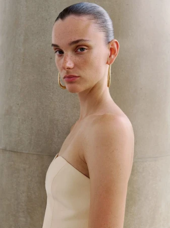 Close-up portrait of a woman wearing a cream strapless top and gold hoop earrings against a neutral pillar background.
