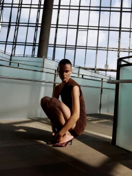 Model crouching in a brown dress and dark heeled shoes inside a modern glass building with a large grid window.