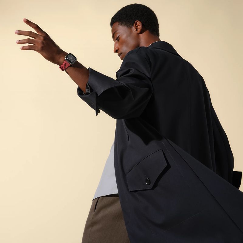 A dark-skinned adult male poses in a black coat and displays a smartwatch on his wrist against a light tan backdrop in a fashion shot.