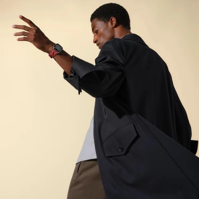 A dark-skinned adult male poses in a black coat and displays a smartwatch on his wrist against a light tan backdrop in a fashion shot.