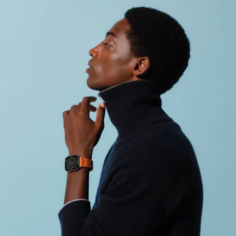 Profile of a dark-skinned man wearing a navy turtleneck and an orange smartwatch on a light blue background in a studio setting.