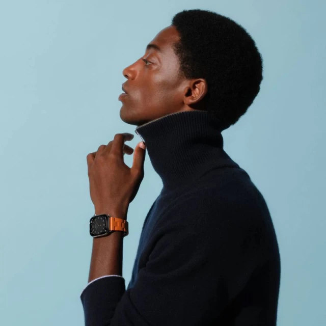 Profile of a dark-skinned man wearing a navy turtleneck and an orange smartwatch on a light blue background in a studio setting.