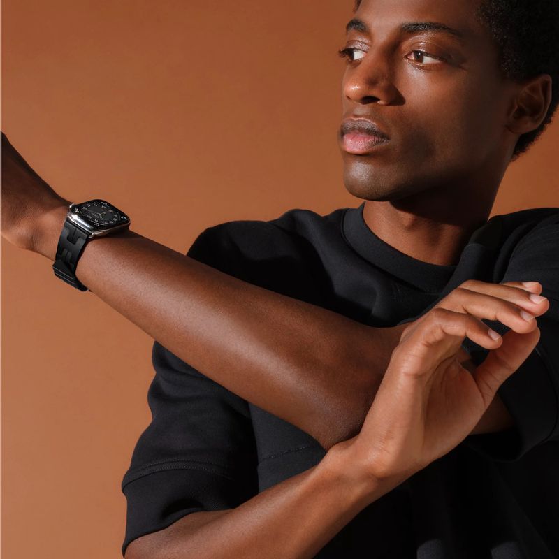 An adult male with dark skin poses with an arm raised wearing a black smartwatch against a neutral brown backdrop in a studio setting.
