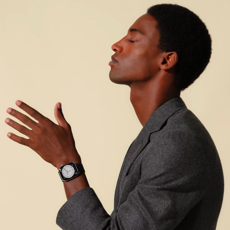 Portrait of an African American male model wearing a grey blazer and a wrist watch, posing against a light yellow background.