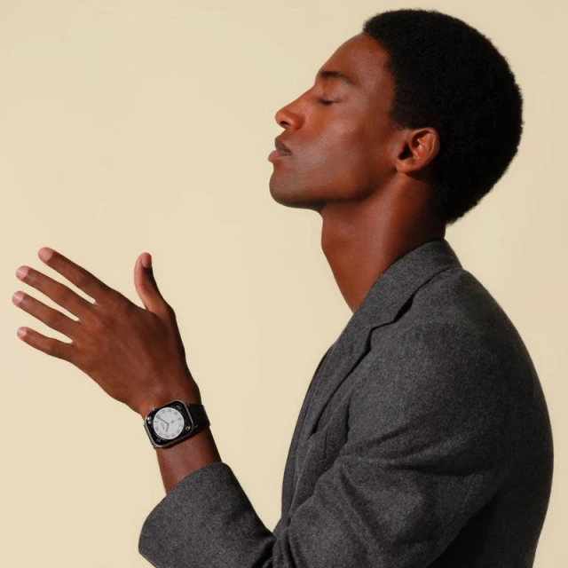 Portrait of an African American male model wearing a grey blazer and a wrist watch, posing against a light yellow background.