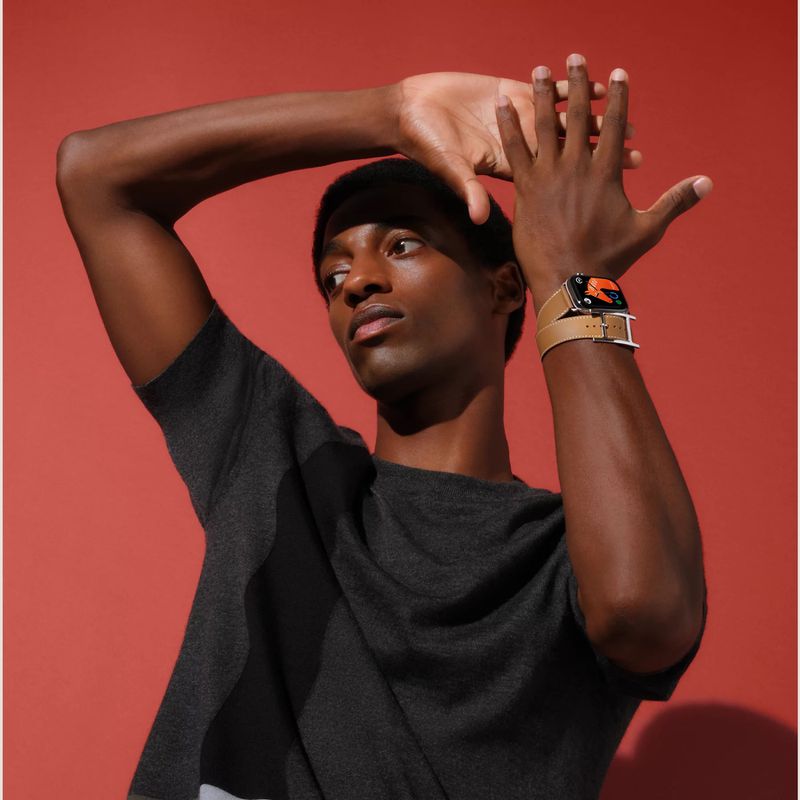 An adult male raises his hands to display a smartwatch with a tan band. He's wearing a gray top against an orange studio backdrop.