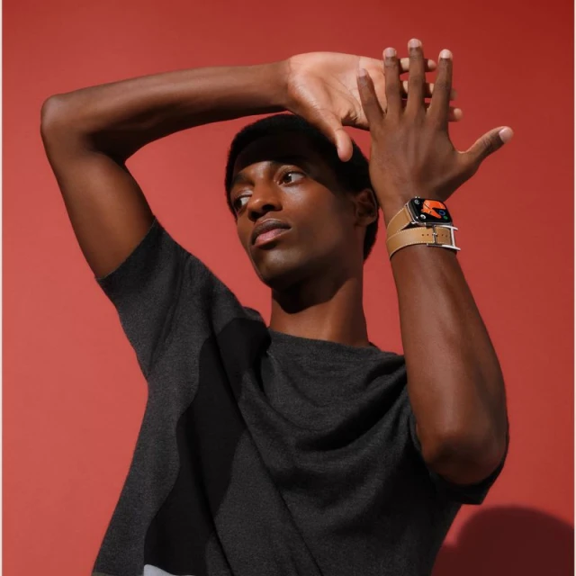 An adult male raises his hands to display a smartwatch with a tan band. He's wearing a gray top against an orange studio backdrop.