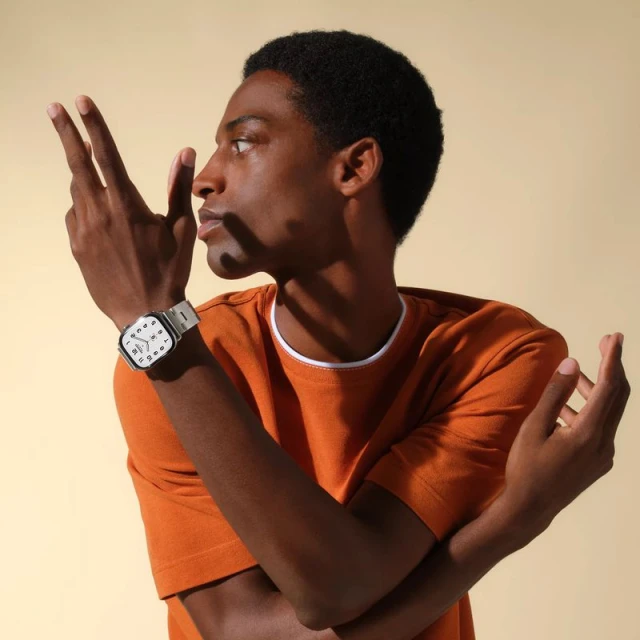 Dark-skinned man in an orange t-shirt poses in front of a tan background wearing a silver wristwatch with a white face.