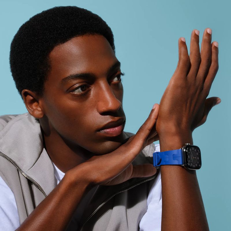 Portrait of a young adult Black man wearing a smart watch with a bright blue wristband against a light blue background.