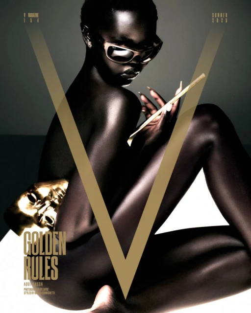 Dark-skinned fashion model posing with a gold mask on a magazine cover, wearing sunglasses and holding sticks.
