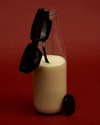 Black sunglasses resting on a glass bottle filled with a creamy white liquid against a dark red background.