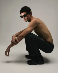 Shirtless man wearing black sunglasses and dark trousers crouching against a grey studio background