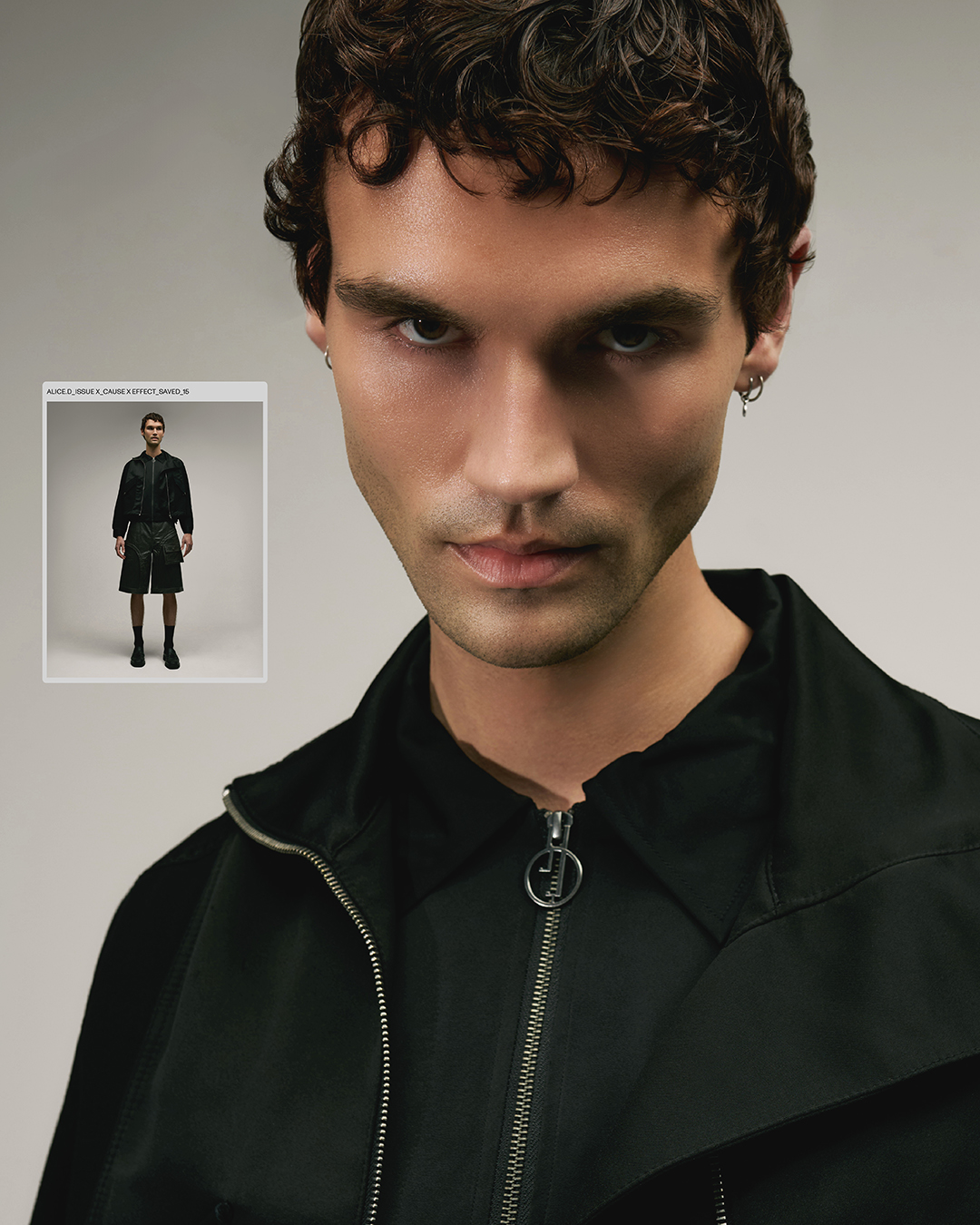 Portrait of a young adult male model with dark curly hair wearing a black jacket, standing indoors against a neutral studio backdrop.