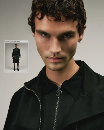 Portrait of a young adult male model with dark curly hair wearing a black jacket, standing indoors against a neutral studio backdrop.