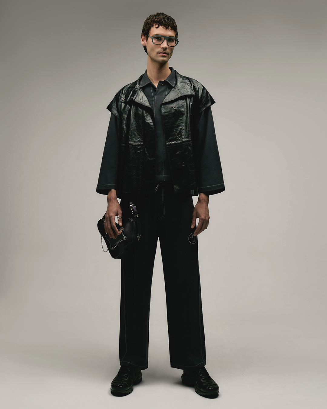 Man wearing a dark jacket and trousers holding a black handbag against a neutral studio background.