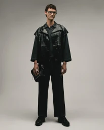Man wearing a dark jacket and trousers holding a black handbag against a neutral studio background.