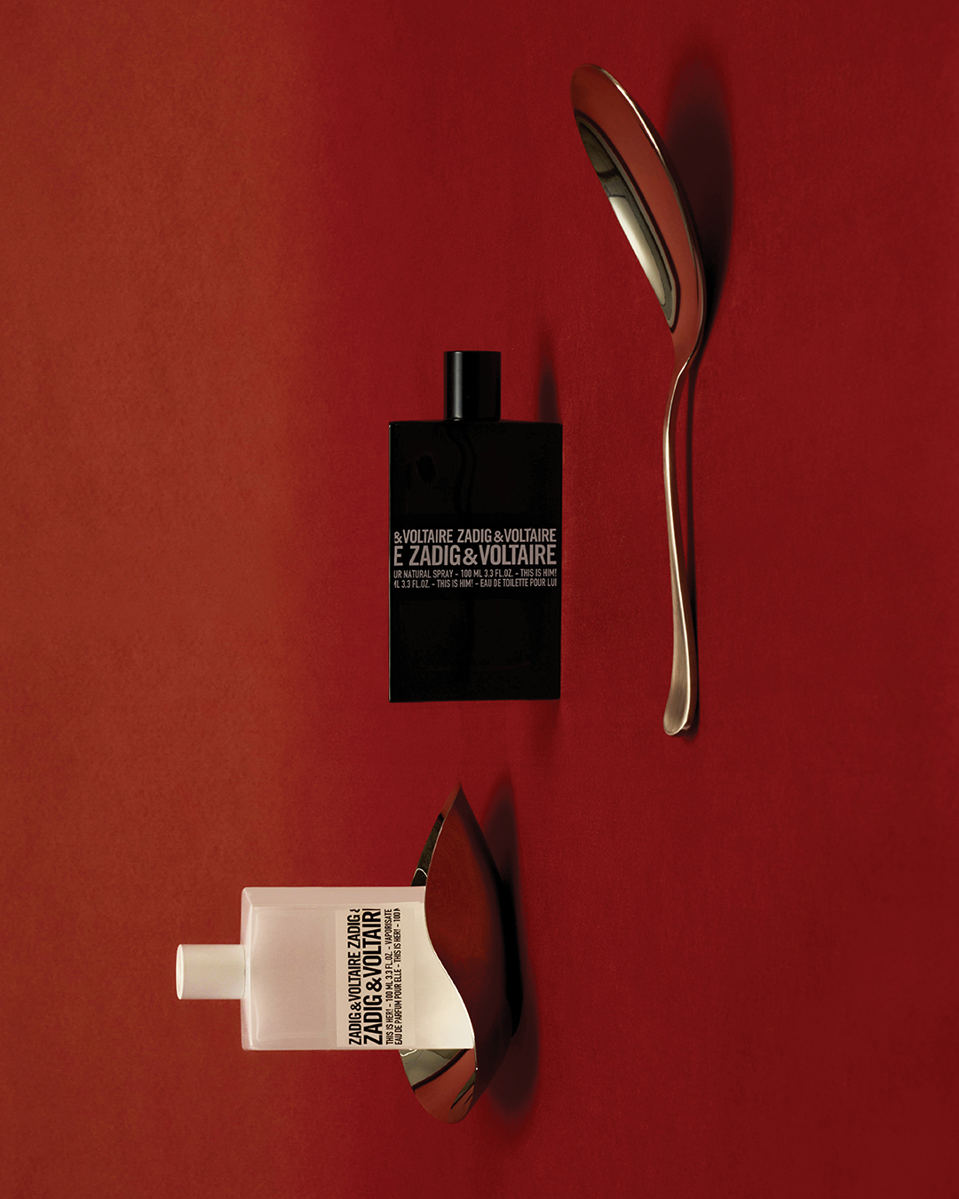 Still life of Zadig & Voltaire perfume bottles, one black, one white, alongside silver spoons on a red background, studio lighting.