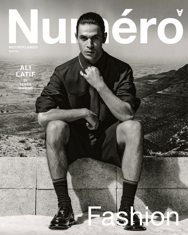 Black and white magazine cover shot of a male model seated on a stone structure with mountains in the background wearing a button-down shirt.