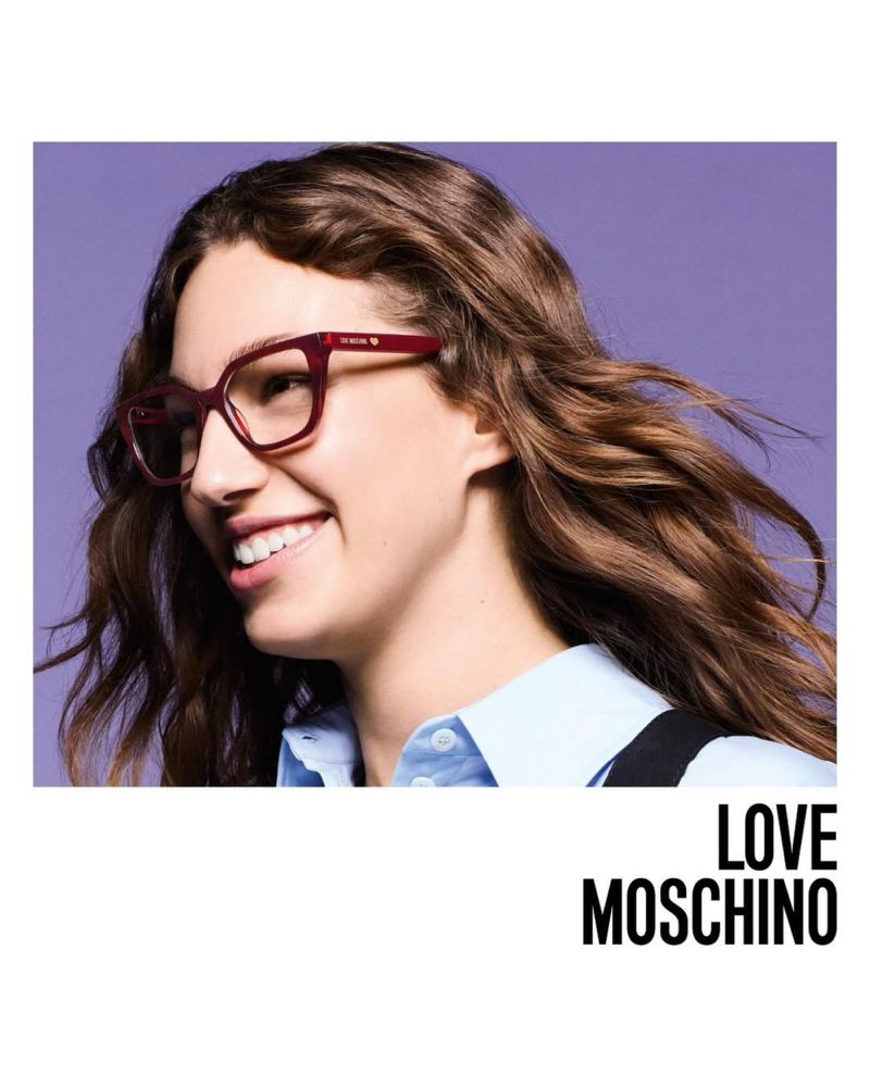 Close-up shot of a smiling woman wearing red eyeglasses, a light blue shirt, and black suspenders against a light purple background.