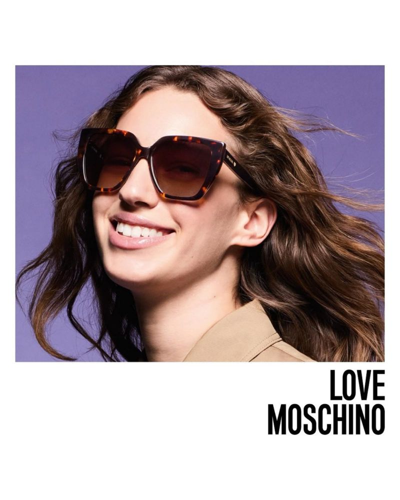 Woman with brown wavy hair smiles wearing brown-tortoiseshell sunglasses and a tan coat in a close-up studio fashion shot.