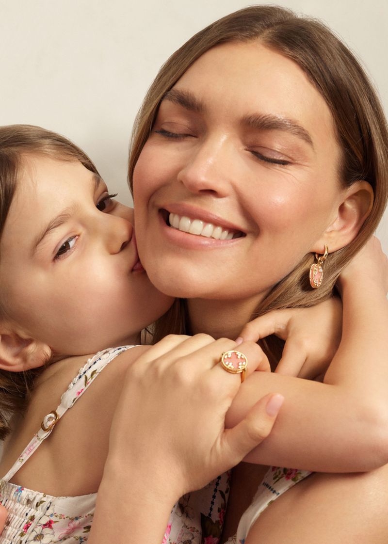 Mother and daughter portrait in a warm, brightly lit setting. The daughter is kissing the mother's cheek; both wear floral-patterned tops.