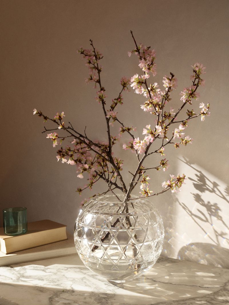 Still life shot of pink blossom branches in a glass vase on a marble surface with a teal glass candle holder and tan book.