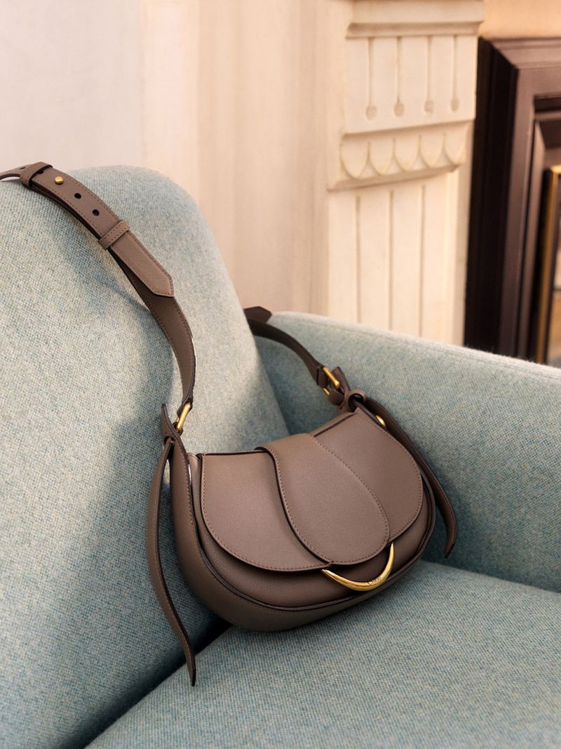 Grey saddlebag purse with a gold clasp, displayed on a teal upholstered chair with decorative molding in the background.