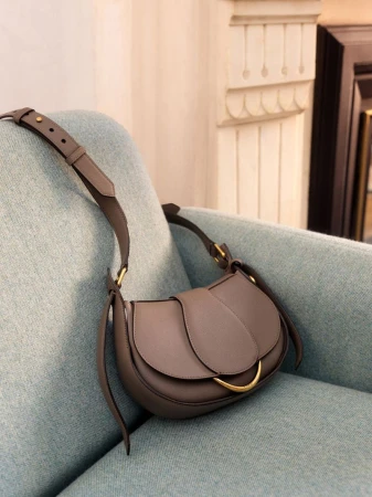 Grey saddlebag purse with a gold clasp, displayed on a teal upholstered chair with decorative molding in the background.
