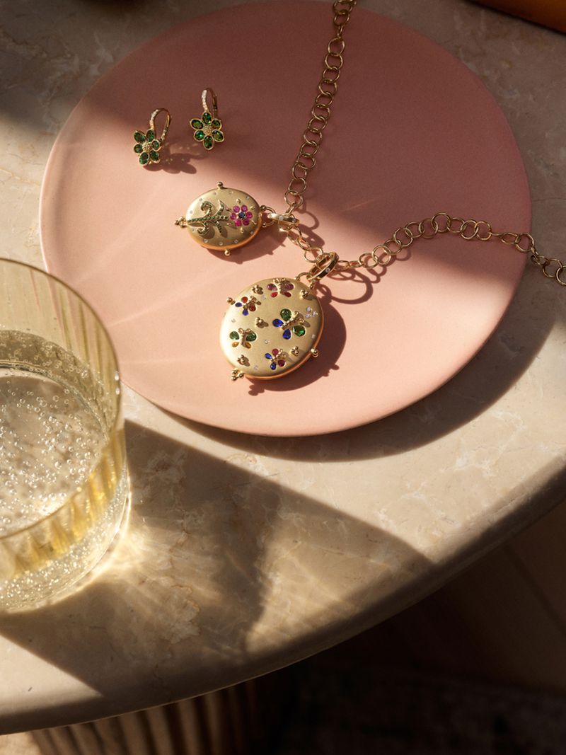 Gold necklace and earrings with floral detail on pink plate beside glass of sparkling wine, lit by warm light on marble surface.
