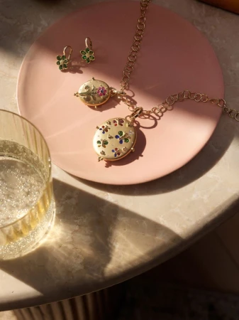 Gold necklace and earrings with floral detail on pink plate beside glass of sparkling wine, lit by warm light on marble surface.