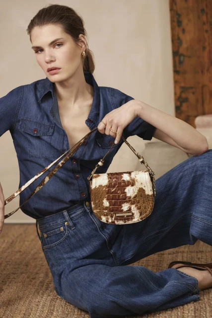 Adult female fashion model sits in a denim outfit holding a brown-and-white Brahmin handbag in a studio shot for fashion advertisement.