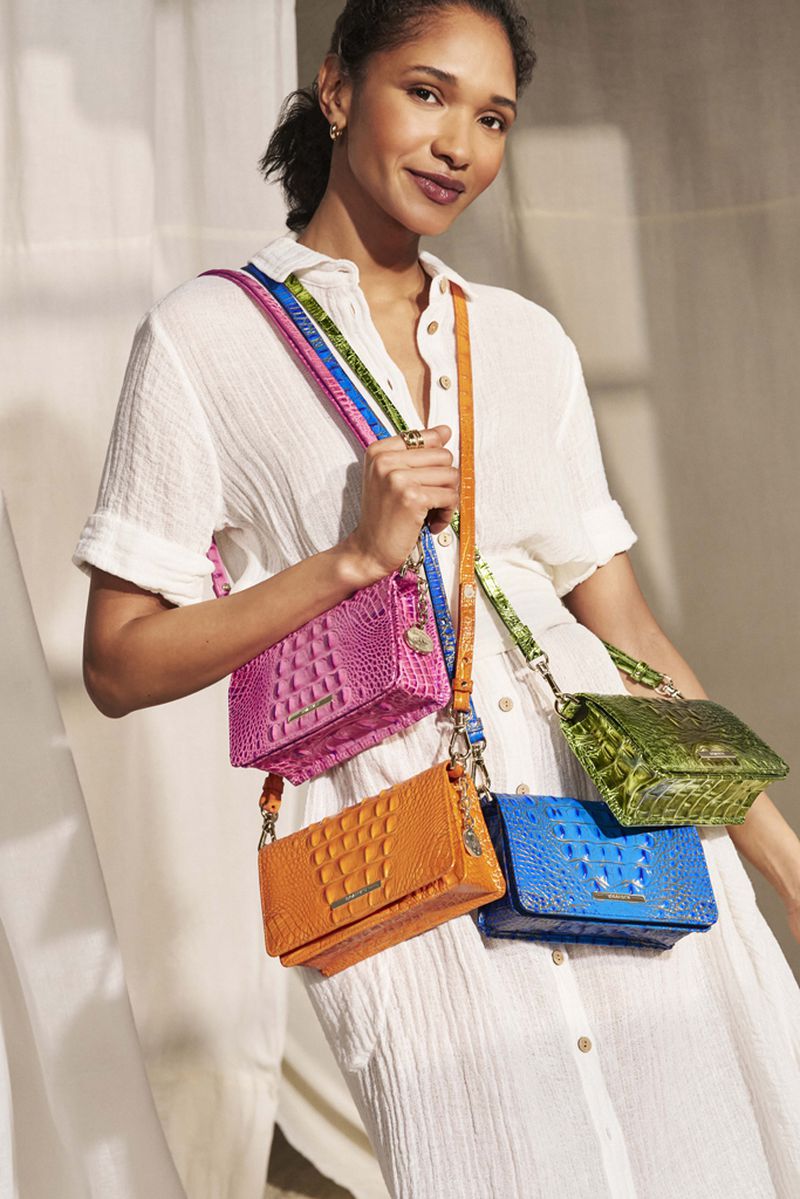 Woman in white dress showcases a collection of colorful crocodile-embossed leather crossbody bags in pink, orange, blue, and green.