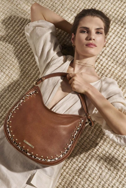 Woman lying on a woven rug holding a brown leather hobo handbag with gold studs and animal print details, wearing a light-colored linen dress.