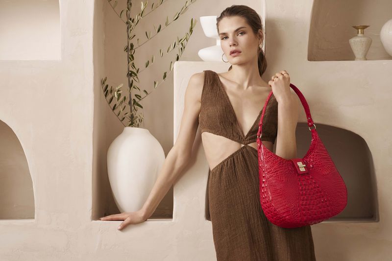 Woman poses with red crocodile-skin handbag in front of white stucco wall with arched niches and vase with leafy branches.