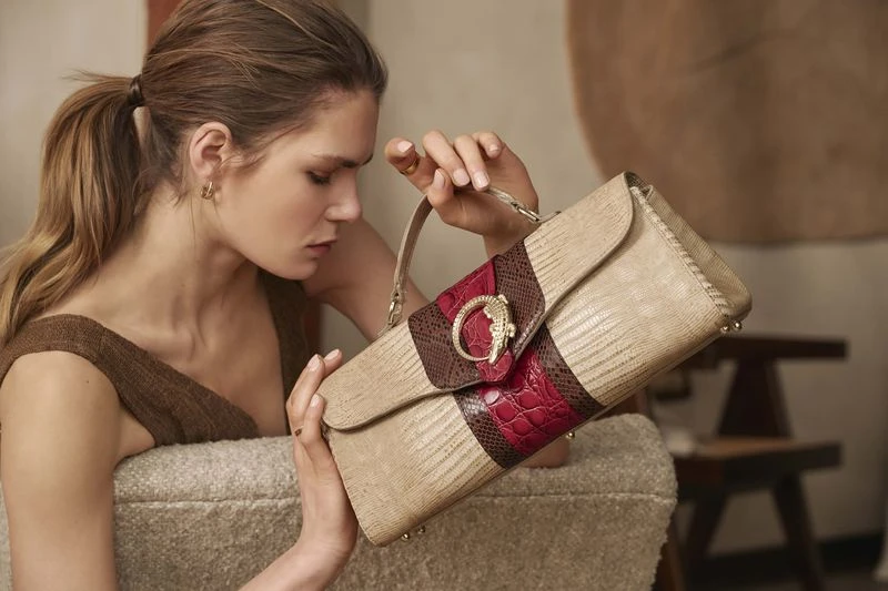 Woman presents a beige, red, and brown handbag with an ornate buckle, featuring reptile skin textures and gold hardware, for fashion editorial.