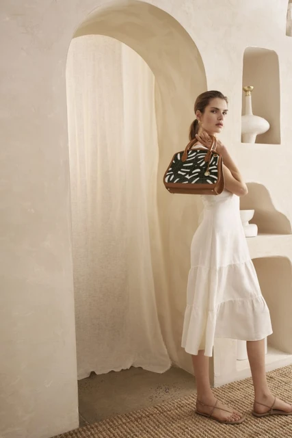 Elegant woman in a white dress poses with a patterned handbag in a sunlit, arched doorway. Neutral tones and natural light.