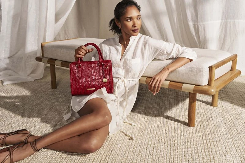 Elegant adult female poses on a textured rug with a red leather satchel handbag, wearing a white dress near a wood bench.