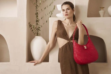 Model wearing a brown cut-out dress holding a red leather shoulder bag against a textured beige architectural wall