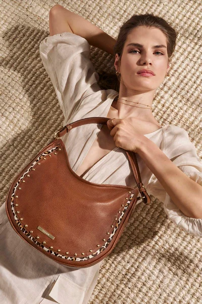 Model lying on a woven rug holding a brown leather shoulder bag with studded trim for Brahmin Summer '25 campaign.