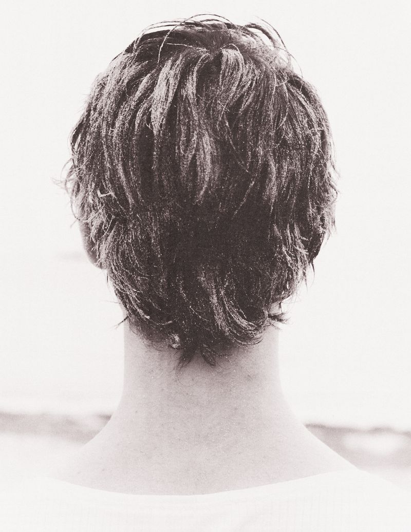 Back of adult human head with short hair, neutral skin tone, wearing a white shirt, against a soft-focus background, monochrome.