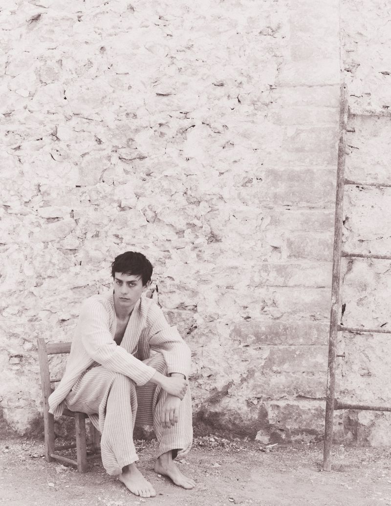 Monochrome portrait of a young adult male sitting on a wooden chair against a stone wall with a ladder, looking contemplative.
