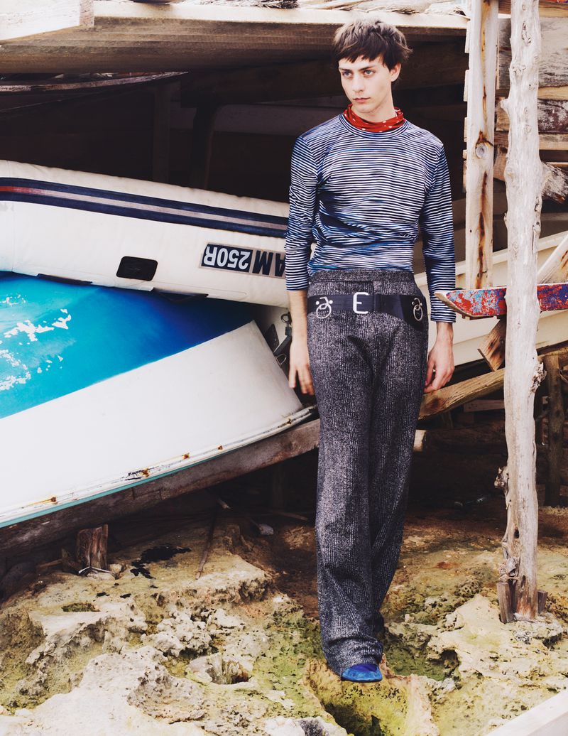 Young male fashion model in striped top and grey tweed pants poses next to boats on a weathered beach location.