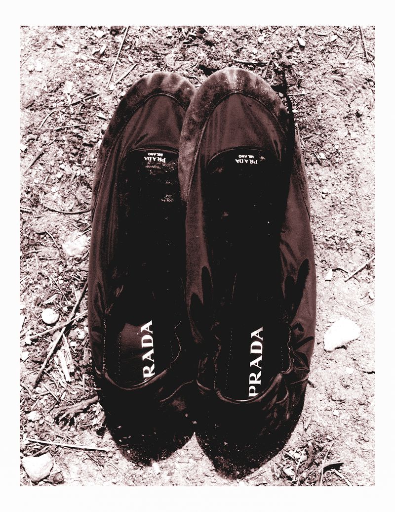 Close-up of a pair of black Prada ballet flats with the brand name visible on the inner sole, sitting on a tan, textured surface.