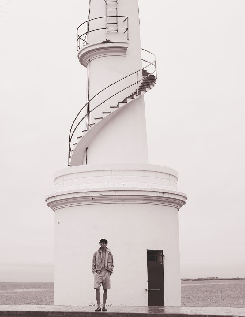 Man standing near a white lighthouse by the sea on an overcast day; wearing a light jacket, shorts, and cap.