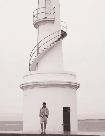 Man standing near a white lighthouse by the sea on an overcast day; wearing a light jacket, shorts, and cap.