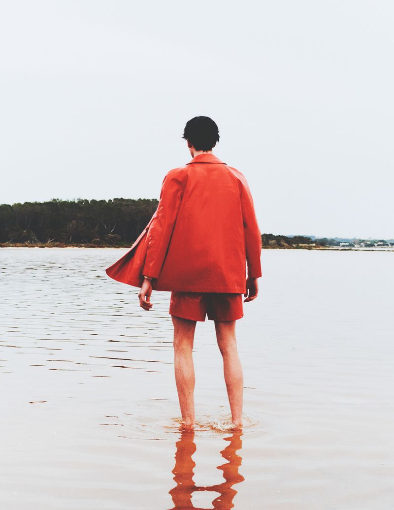 A person in an orange jacket and shorts stands in shallow water near a shore, with a jacket sleeve lifted by the breeze.