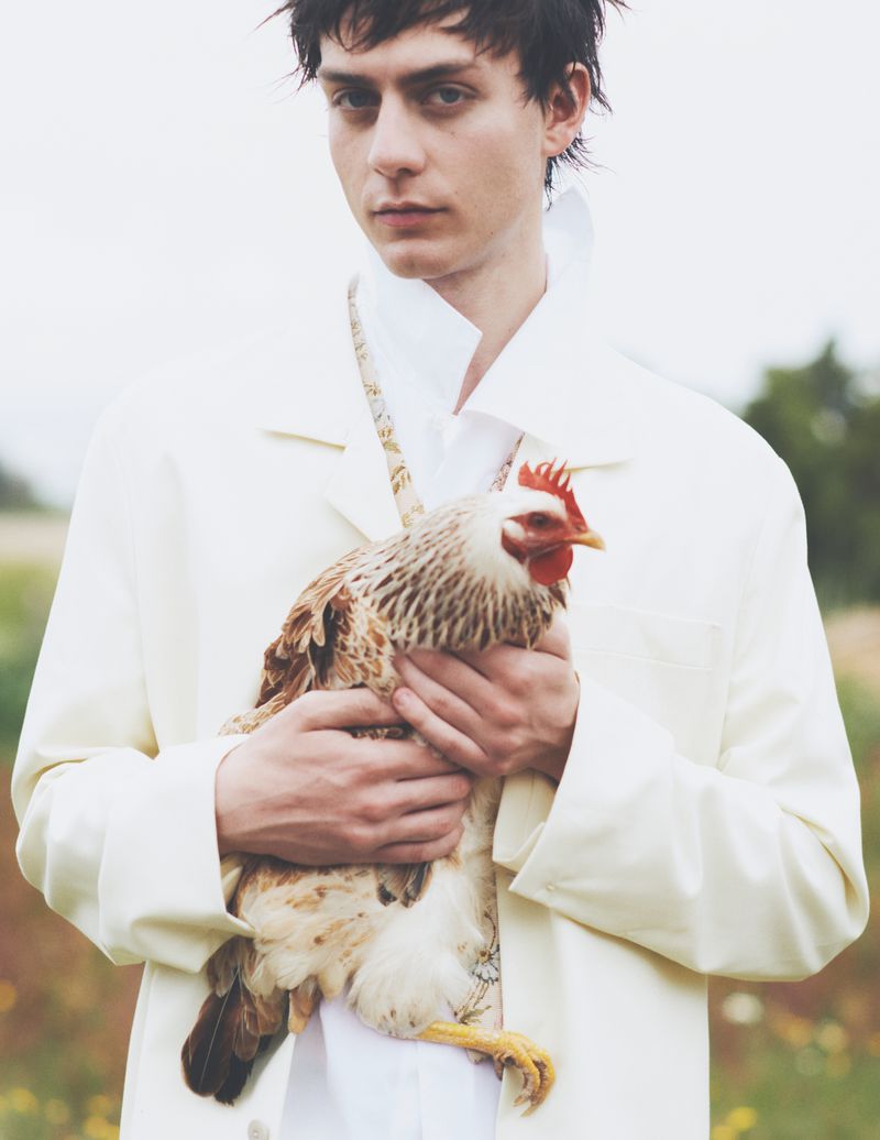 Portrait of a dark-haired young man in a white jacket cradling a hen with mottled feathers in a field on an overcast day.