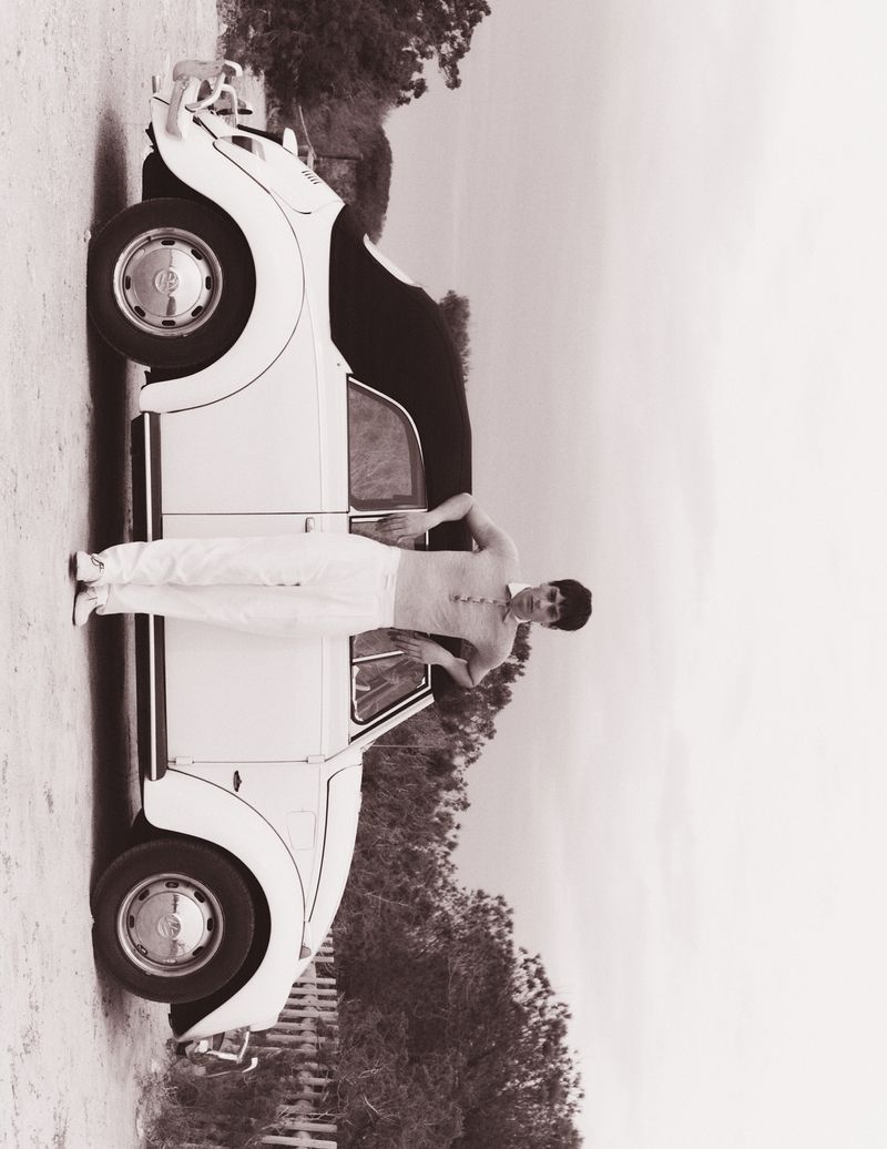 Black and white shot of a female leaning on a Volkswagen Beetle. The setting shows a gravel-covered lot with trees and cloudy skies.