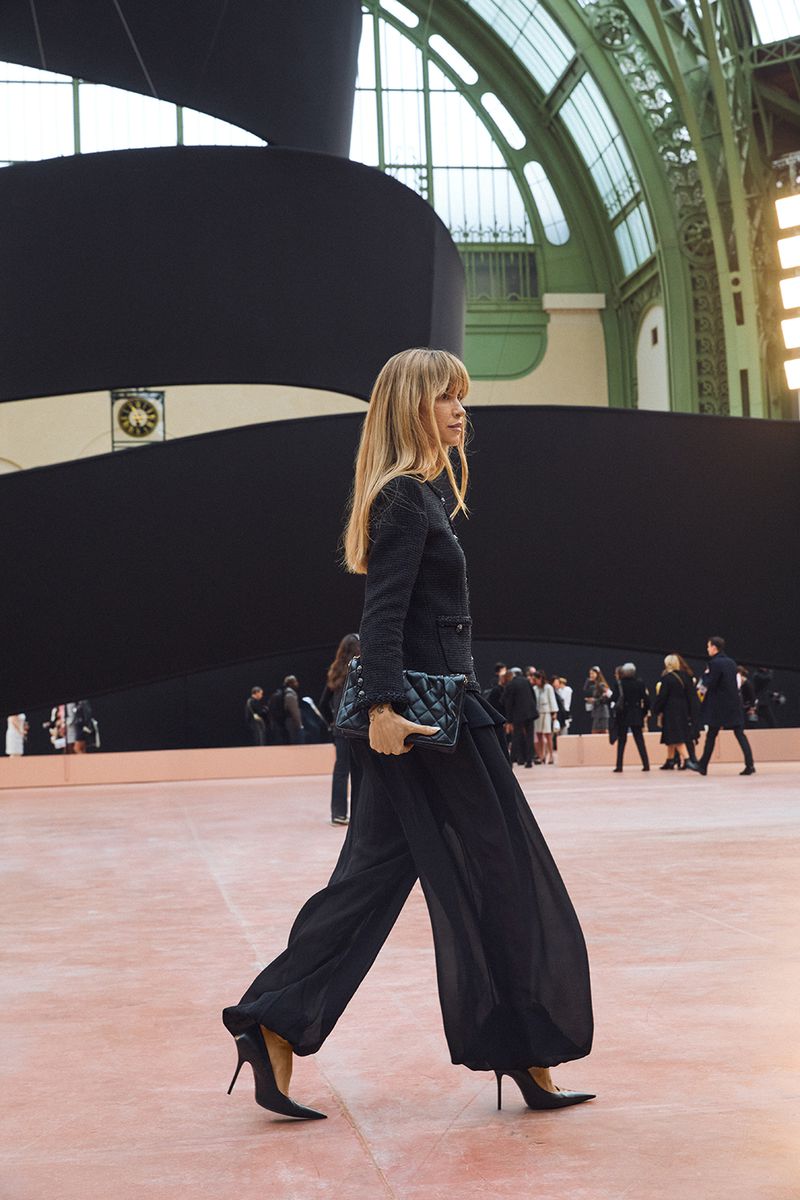 Runway model showcases a dark outfit with black pumps and a matching purse, walking on a light pink catwalk during a fashion show event.